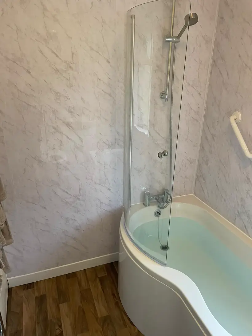 P-shaped shower bath with a glass screen against white marble-effect wall panels, over a wood-effect floor
