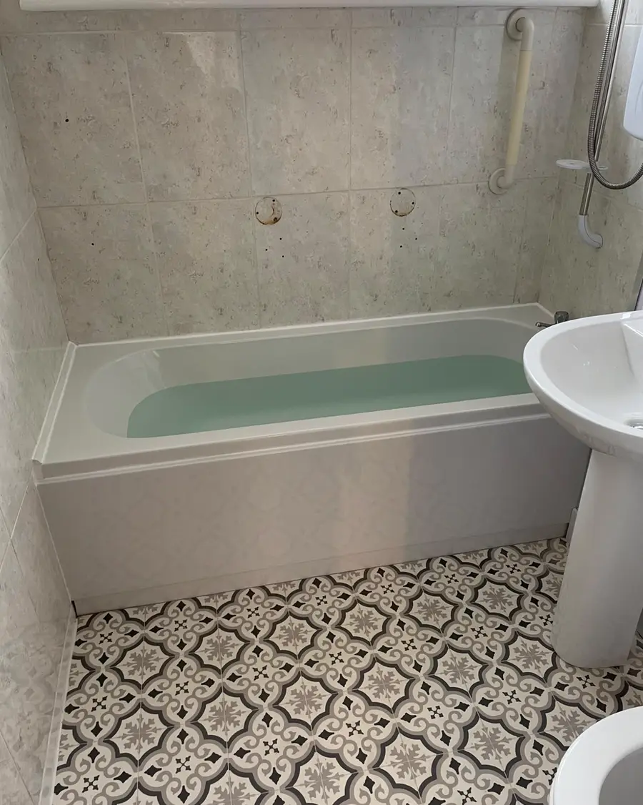 White bath and pedestal basin with marble-effect walls and a patterned tiled floor